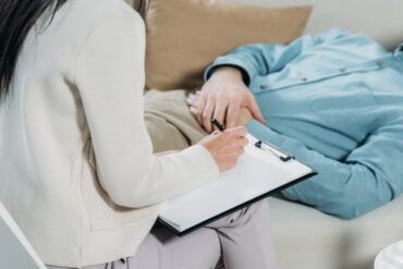 Person lying on a couch while another person takes notes on a clipboard during a therapy session.
