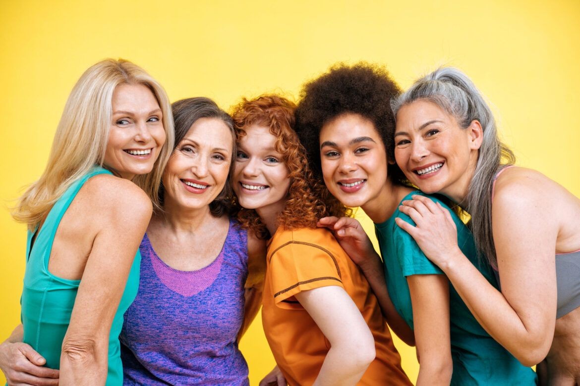 Group of five smiling women standing close together against a bright yellow background.