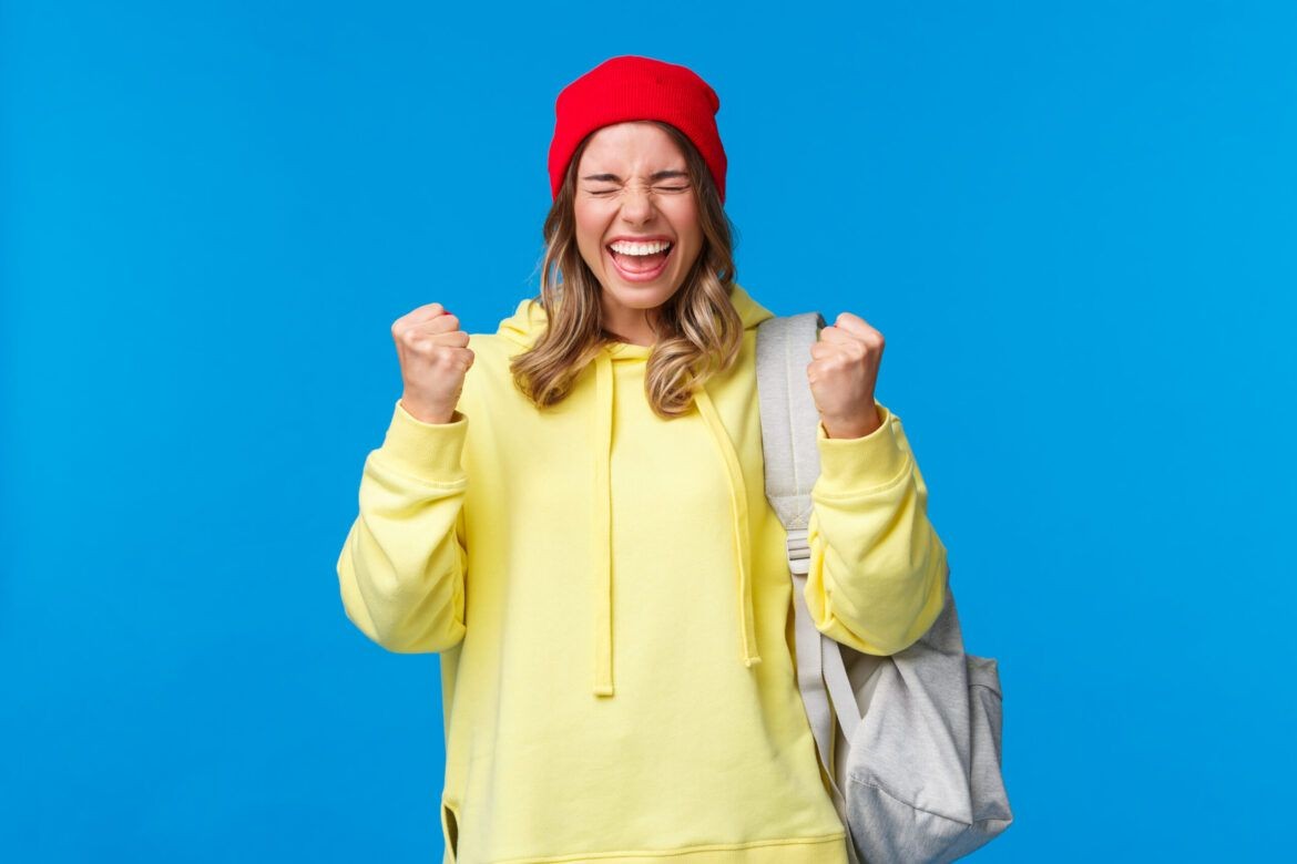 Person in a yellow hoodie and red beanie smiling with clenched fists, against blue background.