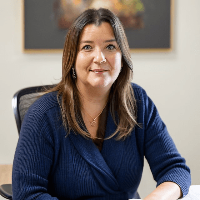 Natalie the hypnotherapist with long hair sitting at a desk, wearing a navy blue sweater and necklace, smiling.