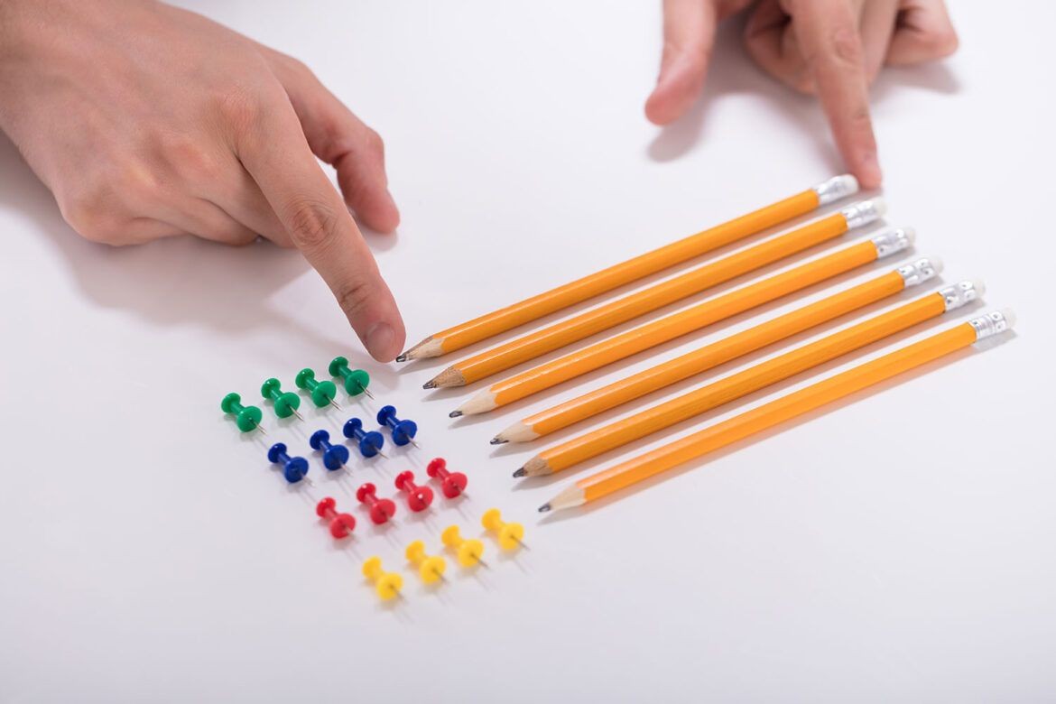 Hands pointing at a row of yellow pencils and colorful push pins on a white surface.