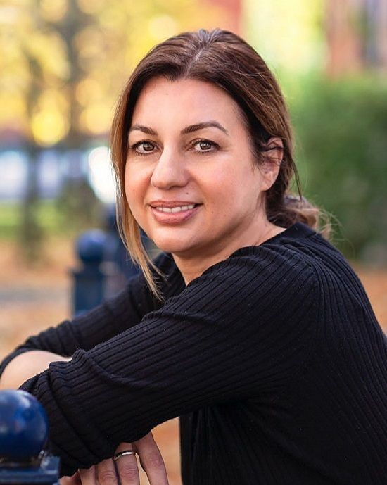 Woman in black sweater smiling outdoors with blurred autumn background.