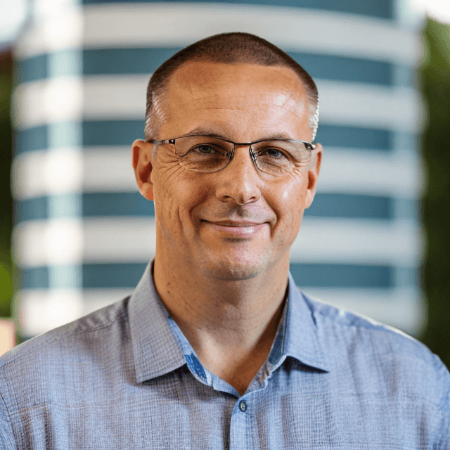 J Lowson wearing glasses and a blue shirt, standing in front of a blurred striped background.