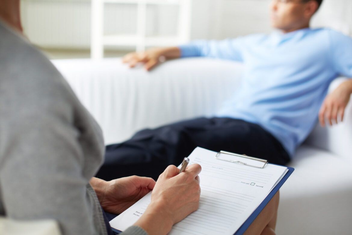 Person writing on a clipboard during a therapy session while another person relaxes on a couch.